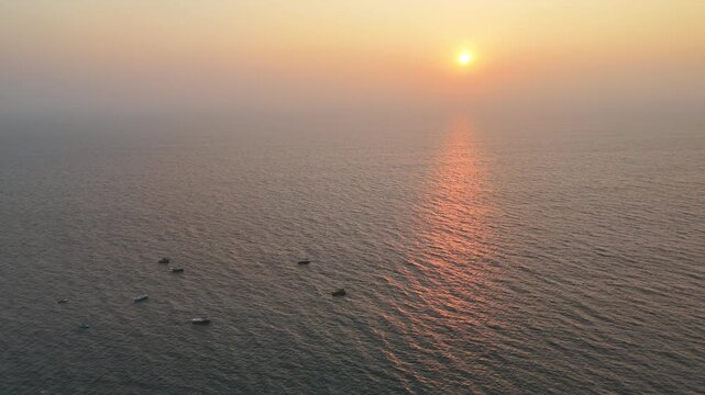 Golden sunset over a calm ocean, with soft waves reflecting warm light across the water&rsquo;s surface. Small boats drift quietly in the distance as the sun lowers into a hazy horizon, creating a peaceful