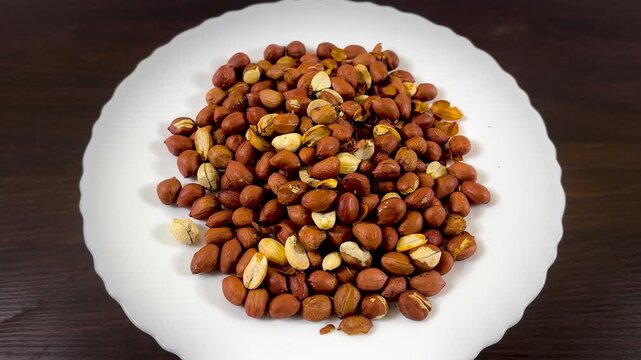 Roasted peanuts with skins on white plate, crunchy salted groundnuts on dark wooden table, healthy protein snack for culinary and diet concepts