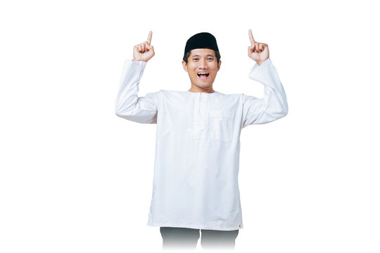 Happy young Muslim man in white baju kurung and black peci pointing both index fingers upward against white background