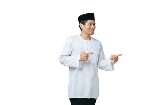 Happy young Muslim man in white baju kurung and black peci pointing both index fingers on his side against white background
