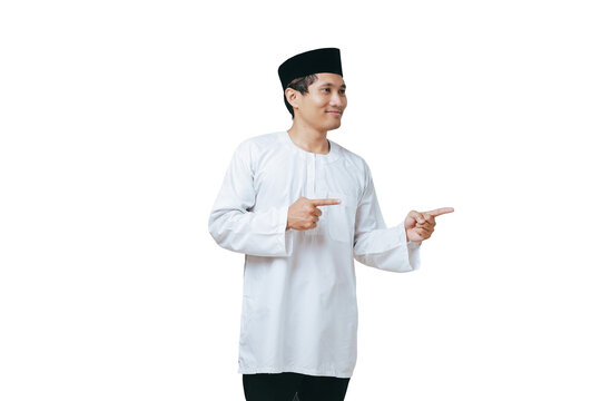 Happy young Muslim man in white baju kurung and black peci pointing both index fingers on his side against white background