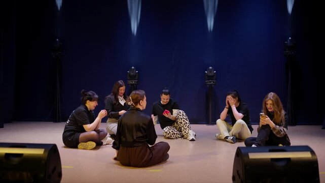 Group of young women sitting in a circle on a dimly lit stage, rehearsing and talking together in a relaxed atmosphere with speakers and spotlights around them