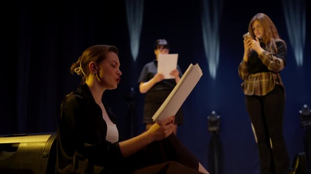 Aspiring actors rehearsing dramatic script on dimly lit theater stage, seated and standing performers practicing lines with papers under focused stage lighting