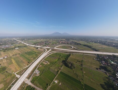 Aerial view of a modern highway interchange surrounded by rice fields and rural settlements with a distant volcano under a clear sky in Klaten, Central Java, Indonesia.
