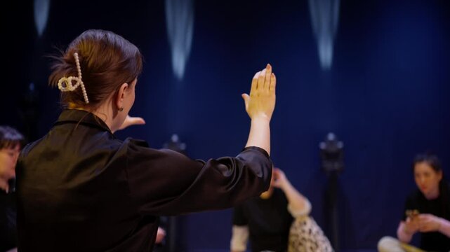 Contemporary theatre rehearsal with ensemble of young performers sitting on stage as expressive director guides movement and gesture under dramatic blue lighting