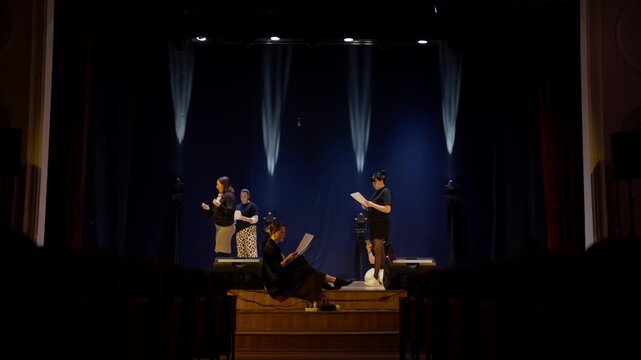 Rehearsal of a dramatic stage performance with actors reading scripts under theatrical lighting on a dark theater stage before a live audience gathering