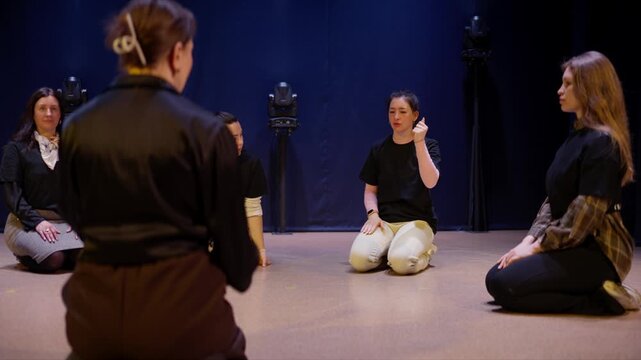 Group of performers rehearsing expressive movement sequence in a dimly lit studio, kneeling in a circle and practicing synchronized theatrical gestures together