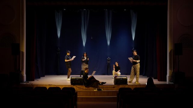 Small theatre rehearsal on a dimly lit stage with actors practicing lines, expressive blocking and performance timing in front of empty audience seats