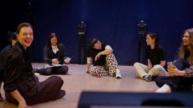 Creative workshop circle with women sharing stories and laughter while seated on the floor in a dimly lit studio rehearsal space with theatrical lighting
