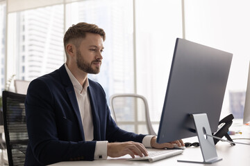 Fototapeta premium Focused young Caucasian businessman in formal suit sits at desk, working on desktop computer in modern office, concentrating on business tasks, online communication. Workflow using tech, productivity