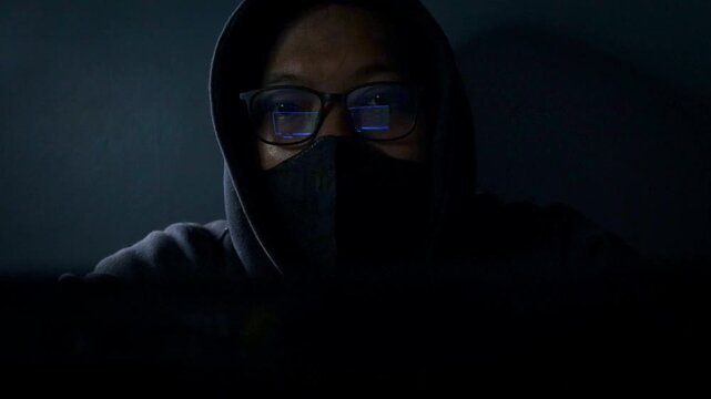 Hacker wearing glasses and face mask conducting cyber attack in a dark room, network breach and ransomware in internet fraud. Night scene with computer, spyware, dark web and online fraud activity.