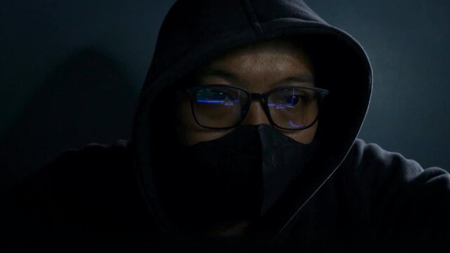 Hacker wearing glasses and face mask conducting cyber attack in a dark room, network breach and ransomware in internet fraud. Night scene with computer, spyware, dark web and online fraud activity.