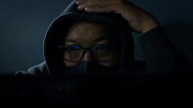 Hacker wearing glasses and face mask conducting cyber attack in a dark room, network breach and ransomware in internet fraud. Night scene with computer, spyware, dark web and online fraud activity.