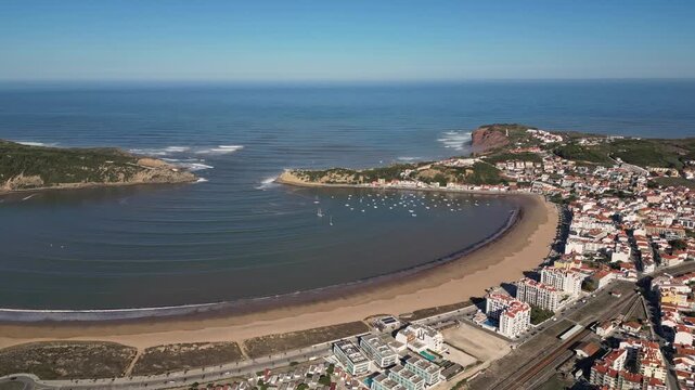 Aerial drone shot panning left with a slight orbit over S&atilde;o Martinho do Porto bay toward Salir do Porto, showing the beach, boats, coastal town and bay entrance in clear morning light.