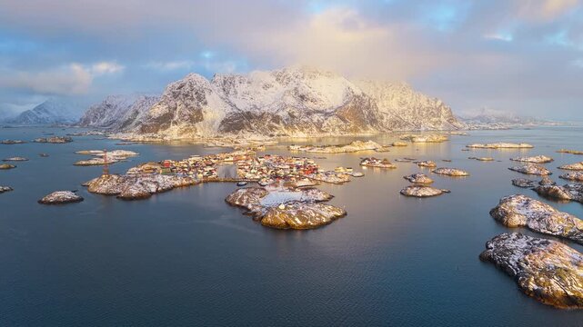 Breathtaking cinematic drone shot of the iconic Henningsvaer fishing village surrounded by snowy mountains and arctic islands during a vibrant winter sunset in Lofoten, Norway.