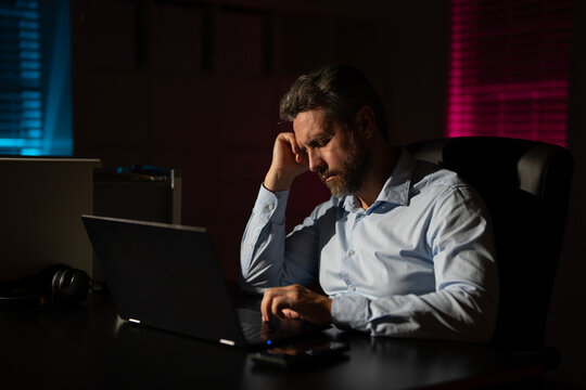 Business man using laptop late at night office. Overworked businessman at a desk with a hard office work. Business man on office night background. Worker bored and tired in dark office.