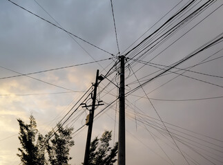 Fototapeta premium Low angle view of utility poles with multiple tangled power lines stretching across a cloudy sky at dusk, highlighting urban infrastructure and energy distribution.