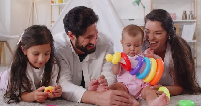 Family with children play educational game with toys at home. Sister and toddler baby explore colorful stacking rings pyramid on floor with parents. Learning, boosting development and leisure. 