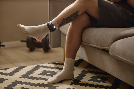Close up of young adult man sitting on sofa with electronic ankle monitor, suggesting house arrest, legal restriction, home confinement. Useful for crime, justice, surveillance campaigns