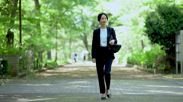 新緑の公園をビジネスバッグを持って歩く女性の営業マンの全身ポートレート仕事イメージの動画