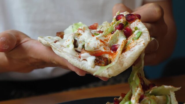 Closeup of a person taking a fresh pita bread sandwich filled with lettuce, pomegranate seeds, and white sauce from a dark plate, ready to eat a healthy and delicious homemade meal