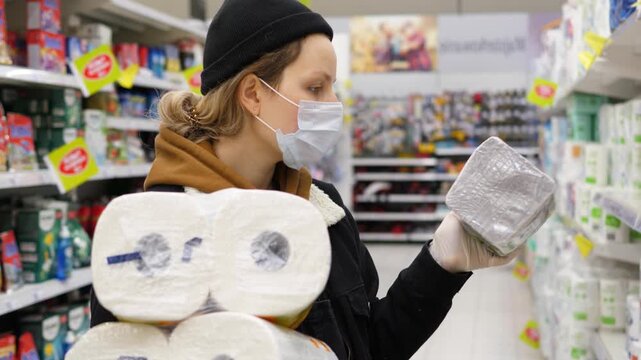 Careful woman wearing a protective face mask and disposable gloves while stocking up on toilet paper in a supermarket aisle, demonstrating consumer behavior during a public health crisis