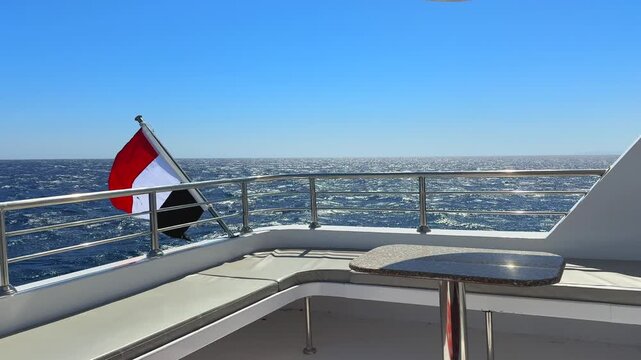 Empty upper yacht deck fenced safety stainless railing and Egyptian national flag behind. Excursion boats stop on the coral reef