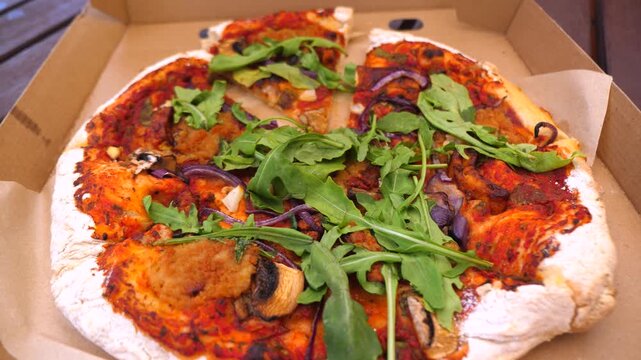 Freshly baked vegan pizza with tomato sauce, mushrooms, red onions, and arugula disappearing slice by slice from a cardboard delivery box, representing healthy eating and takeout food