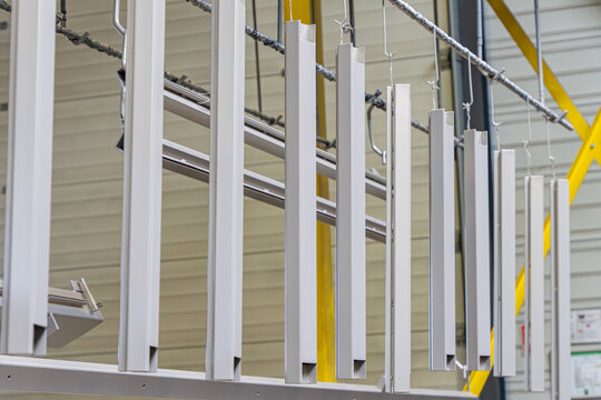 Vue align&eacute;e de profil&eacute;s m&eacute;talliques blancs suspendus verticalement &agrave; un rail de convoyage automatis&eacute; pour le traitement de surface en usine.