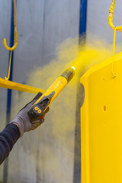 Ouvrier utilisant un pistolet &eacute;lectrostatique pour appliquer un rev&ecirc;tement en poudre jaune vif sur une pi&egrave;ce m&eacute;tallique suspendue dans un atelier industriel.