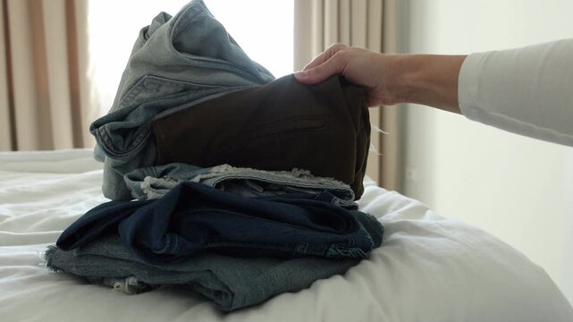 Woman's Hand Picking Folded Denim Jeans from Neat Stack on White Bed &mdash; Sorting Multiple Blue and Gray Shades of Casual Pants Indoors, Wardrobe Organization, Capsule Closet Decluttering and Mindful	