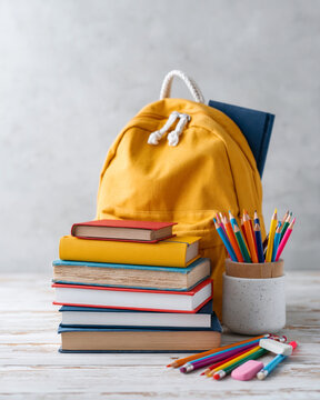 Yellow backpack leans behind stacked books. Pencils rest in a mug beside scattered crayons. Brightly colored volumes suggest learning or creativity. Scene evokes energy and academic readiness