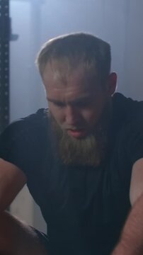 Focused bearded athlete training on indoor rowing machine in dimly lit gym, highlighting endurance workout intensity, effort, and determination during cardio exercise