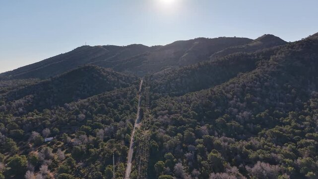 Drone shot following a service road and power infrastructure through the rugged, pine-covered terrain of the Serra Gelada mountains near Benidorm.