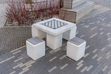 High angle view of concrete outdoor chess table and stools in modern city park. Square concrete game table with an integrated checkered board for chess or checkers, surrounded by three matching stools © Alena