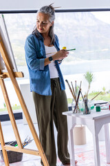 Naklejka premium Senior woman standing at wood easel, holding brush and jar, painting on canvas in coastal studio