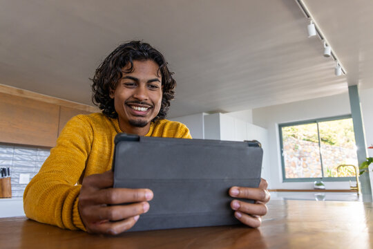 Indian man sitting at kitchen wooden table holding tablet in folio case, wearing mustard sweater
