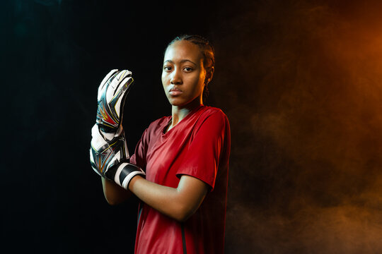 Female athlete holding goalkeeper gloves, in red jersey, studio-lit with teal-orange smoke