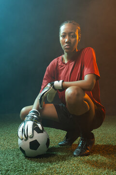 African American woman goalkeeper crouching on turf, stabilizing with gloved hand on soccer ball