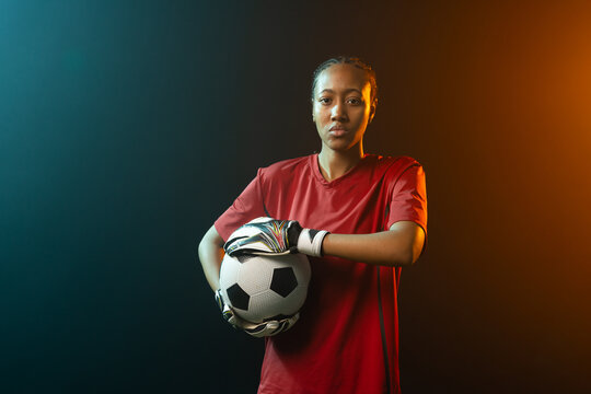 Female goalkeeper holding soccer ball on chest in studio with red jersey gloves teal-orange light