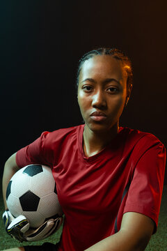African American female keeper posing in red kit, gripping b and w ball, glove, turf, blue-amber lig