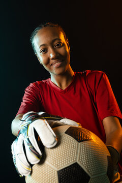 African American female goalkeeper holding soccer ball in studio, wearing red jersey and gloves
