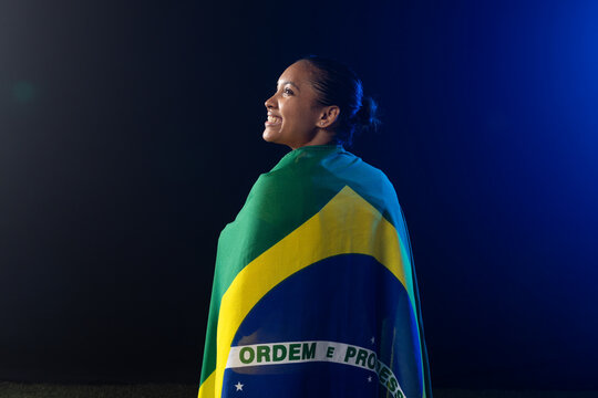 Woman standing wrapped in Brazilian flag showing ORDEM E PROGRESSO, studio blue rim light, grass