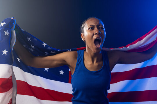 African American woman shouting, holding American flag like cape, wearing blue tank top and studs