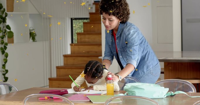 Leaning mother guiding daughter writing with green pencil on wooden dining table, denim shirt