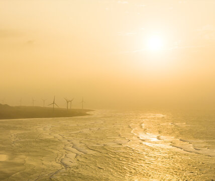 Aerial view of wind turbines along a hazy coastline with waves crashing on the shore during a golden sunset Miaoli County, Taiwan.