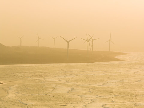 Aerial view of wind turbines standing along a misty coastline with waves crashing on the shore during a golden sunset Miaoli County, Taiwan.