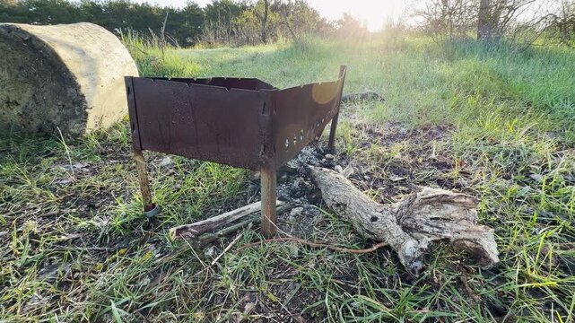 An old metal grill stands ready for a campfire in a quiet, sunlit meadow. The warm morning glow creates a nostalgic and lonely feeling. It is a peaceful spot for a rustic outdoor adventure.