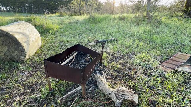 A rusty grill sits quietly in a dew-covered field as the sun rises. This peaceful scene feels calm and nostalgic for outdoor adventures. The soft morning light creates a warm and inviting mood.