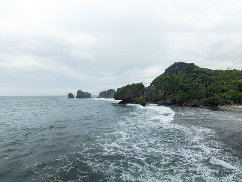 Aerial view of Siung Beach with its iconic limestone cliffs, lush green hills, and white waves crashing against the shore under an overcast sky Tepus, Special Region of Yogyakarta, Indonesia.